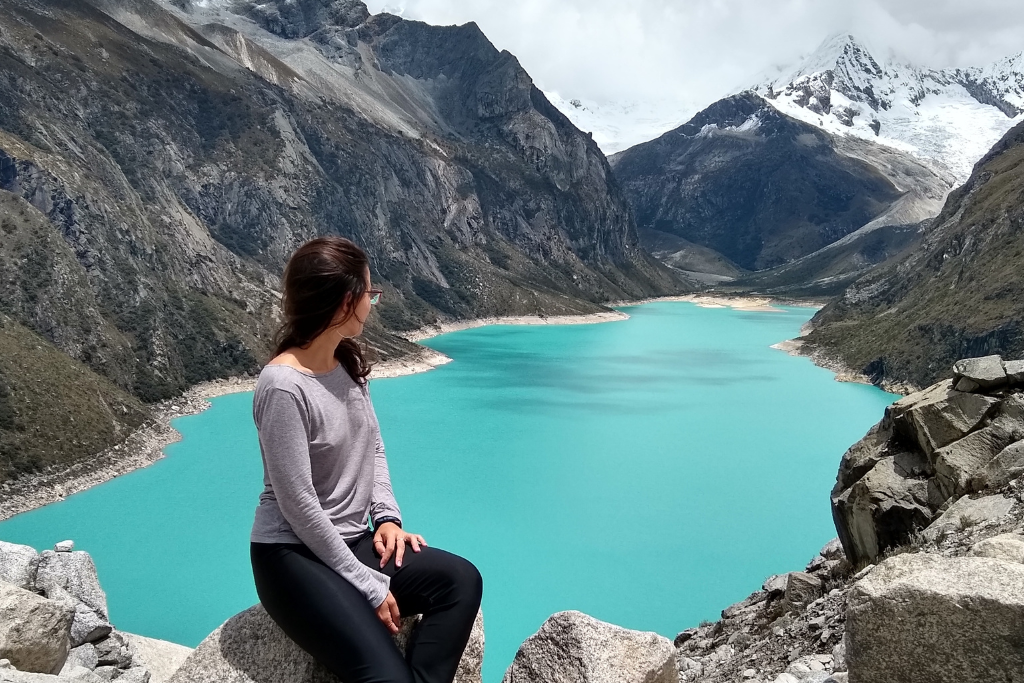 Una mujer sentada sobre rocas grises mira hacia una vasta laguna de color turquesa intenso que llena el centro de un valle profundo. A los lados se elevan montañas escarpadas de granito y, al fondo, se aprecian picos glaciares cubiertos de nieve bajo un cielo con nubes blancas.
