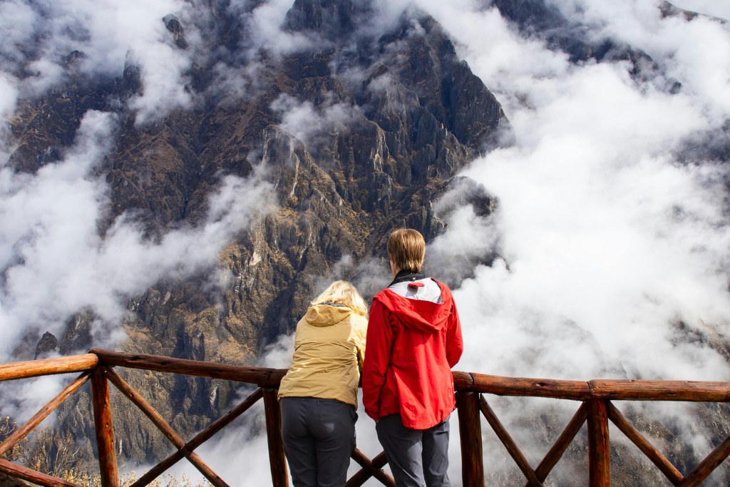 Dos personas de espaldas, una con chaqueta roja y otra amarilla, se apoyan en una baranda de madera para mirar un paisaje de montañas oscuras y afiladas parcialmente cubiertas por densas nubes blancas en el Cañón del Colca.