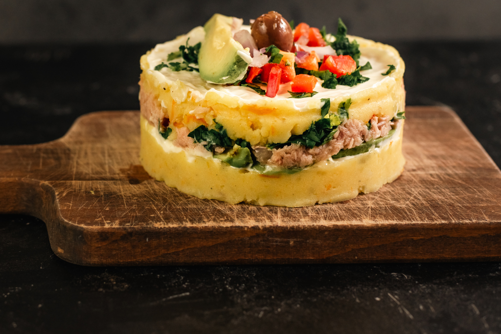 Un pastel redondo de puré de papa amarillo con capas visibles de atún y palta en el centro. El plato está decorado en la parte superior con una rodaja de palta, una aceituna negra, pimiento rojo picado y perejil, presentado sobre una tabla de picar de madera oscura.