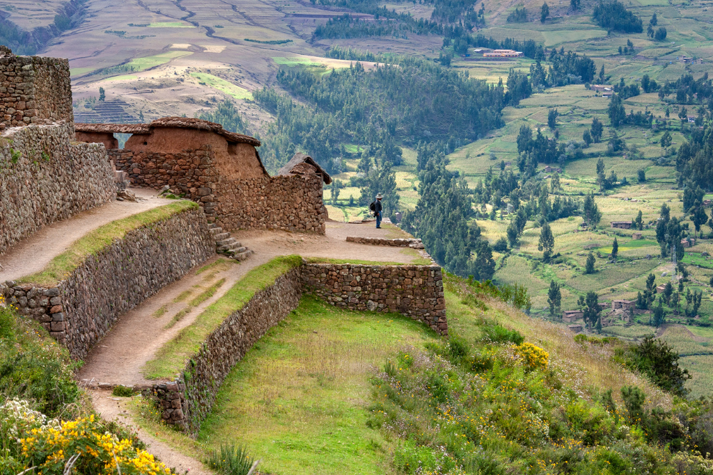 Vista desde lo alto de ruinas incas de piedra y adobe situadas sobre terrazas agrícolas verdes. Una persona contempla el vasto Valle Sagrado al fondo, con sus campos de cultivo y montañas escarpadas bajo una luz suave.