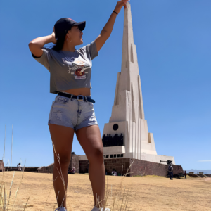 Una joven con gorra negra y pantalones cortos de mezclilla posa de perfil levantando la mano para simular que toca la punta del gran obelisco blanco de la Pampa de la Quinua en Ayacucho