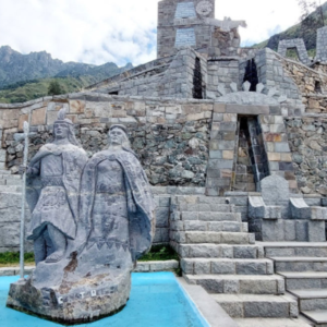 Escultura de piedra gris que representa a la pareja imperial inca, Manco Cápac sosteniendo un cetro y Mama Ocllo a su lado, ubicada sobre una pileta de agua azul brillante. Detrás de las estatuas se alza una imponente estructura de andenes y muros de piedra rústica con escalinatas que conducen a una portada de piedra con un sol tallado en la parte superior y una torre al fondo, bajo un cielo nublado y montañas verdes.