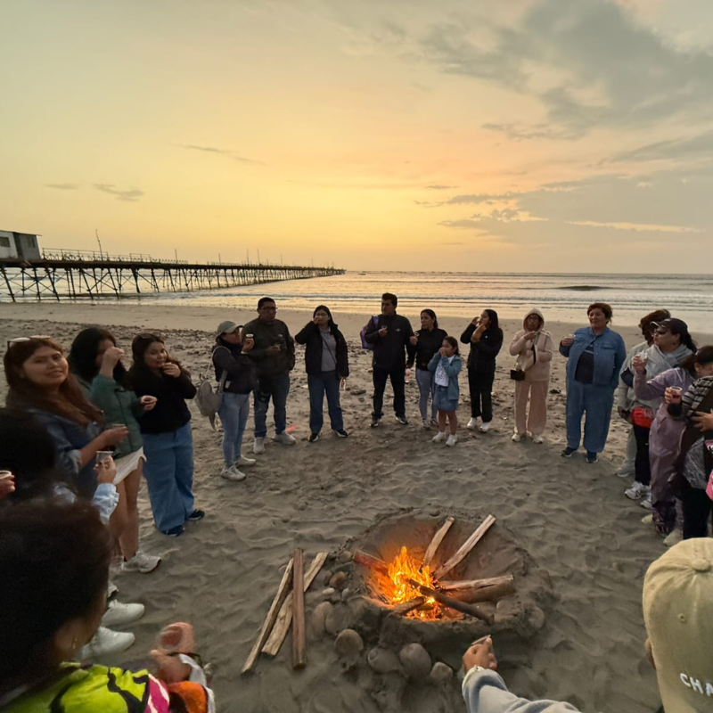 Grupo de turistas reunidos alrededor de una fogata en la playa de Pacasmayo con el histórico muelle de fondo durante el atardecer