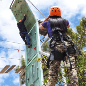 Una joven con casco naranja y arnés de seguridad observa de espaldas a un compañero que asciende por una torre de escalada de madera al aire libre, bajo un cielo azul con nubes en Ayacucho, Perú.