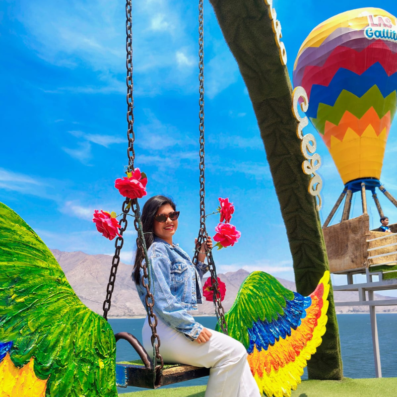 Turista sonriendo en un columpio decorado con flores frente al embalse de Gallito Ciego y un globo aerostático artificial