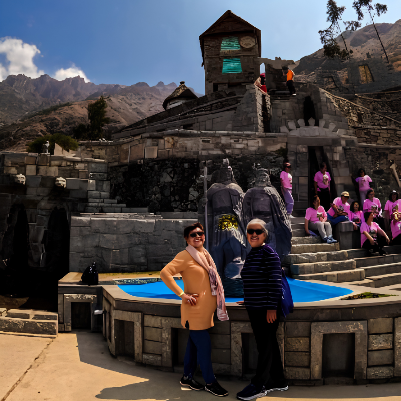 Dos mujeres con gafas de sol sonríen y posan de pie junto a una estructura circular de piedra que contiene una pileta de agua azul brillante. Detrás de ellas se aprecian dos estatuas de guerreros de piedra gris y una gran construcción escalonada con muros y andenes de piedra de estilo incaico. A la derecha, un grupo de personas con camisetas rosadas descansa en las escalinatas, mientras que de fondo se alzan montañas áridas bajo un cielo azul con nubes ligeras