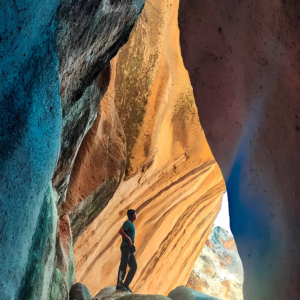 Un hombre de perfil de pie dentro de una cueva o grieta profunda observa las imponentes formaciones de un cañón, donde las paredes rocosas contrastan entre tonos fríos oscuros en primer plano y un fondo fuertemente iluminado en tonos amarillos y ocres.