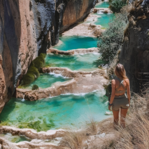 Una mujer rubia vestida con ropa deportiva contempla de espaldas un cañón profundo donde fluye una cadena de pozas naturales escalonadas con agua de color turquesa intenso y formaciones rocosas calcáreas en Millpu, Ayacucho, Perú.