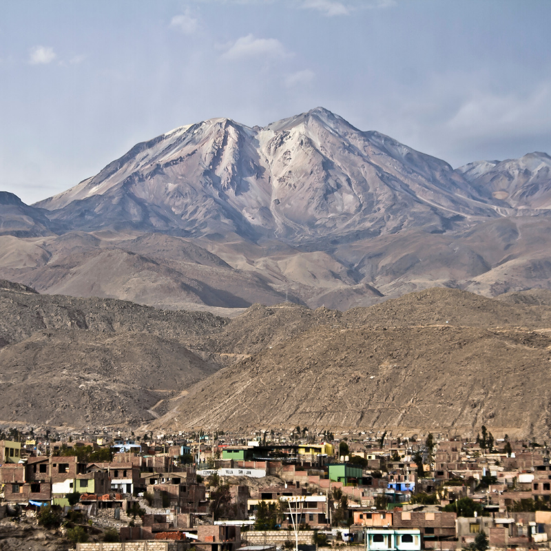 El volcán Chachani con escasa nieve en sus cumbres rocosas, elevándose sobre un denso asentamiento urbano de casas de ladrillo bajo un cielo semicubierto en Arequipa