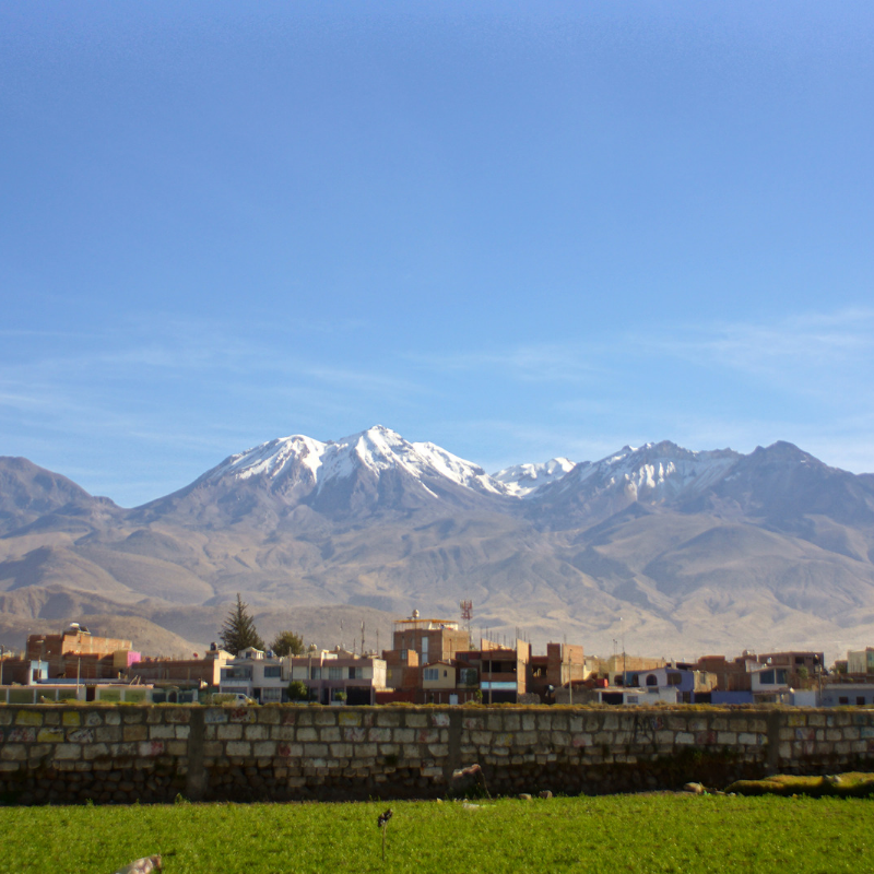 Vista del volcán Chachani con cumbres nevadas detrás de un área urbana y un campo verde en primer plano bajo un cielo azul despejado en Arequipa.