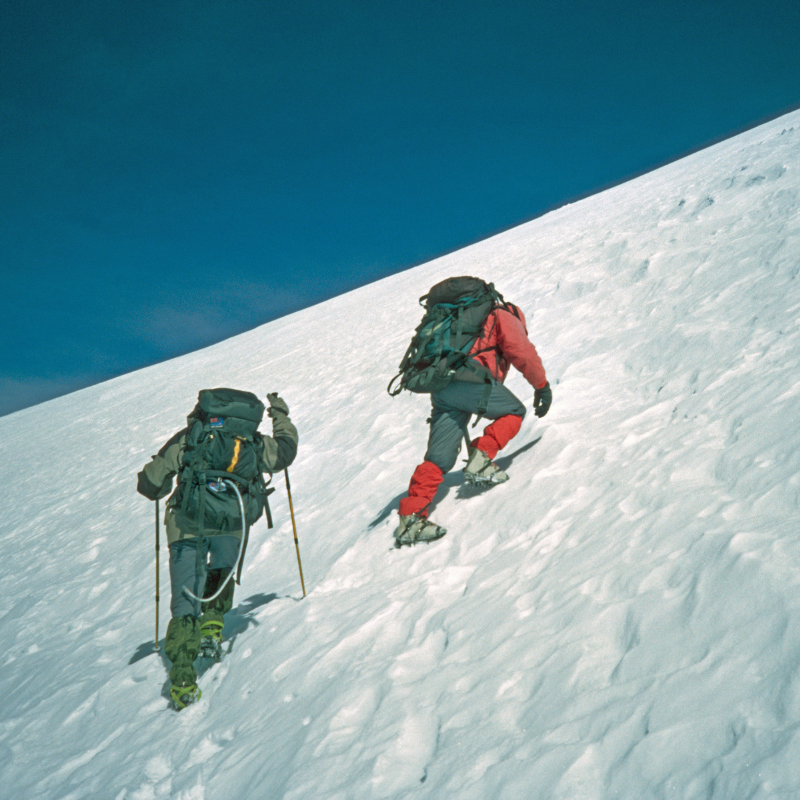 Dos andinistas con mochilas de expedición y bastones de trekking ascienden por una empinada ladera cubierta de nieve bajo un cielo azul despejado durante el tour al volcán Chachani en Arequipa