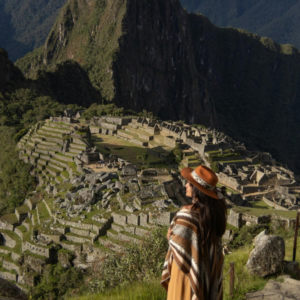Una mujer de espaldas con sombrero marrón y un poncho andino de rayas contempla la antigua ciudadela de Machu Picchu y sus terrazas agrícolas de piedra desde un mirador alto