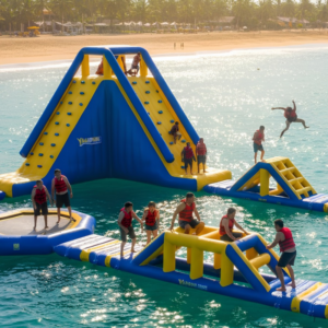 Un grupo de personas con chalecos salvavidas disfruta de un gran circuito inflable de color azul y amarillo flotando en el agua, con toboganes, escaleras y zonas de salto, bajo la luz del sol