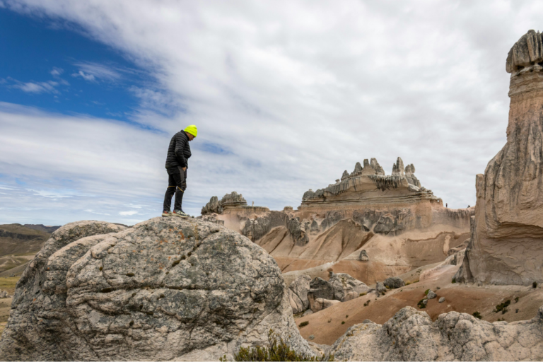 Un viajero con casaca negra y chullo amarillo fluorescente se encuentra de pie sobre una gran roca, contemplando las gigantescas formaciones geológicas de sillar y ceniza volcánica del Bosque de Piedras de Choqolaqa bajo un cielo parcialmente nublado.