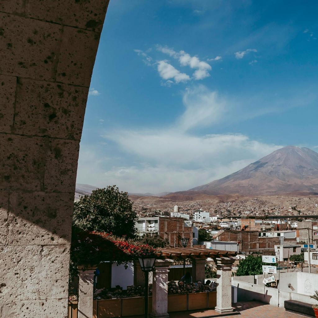 Vista parcial en primer plano de un arco de sillar del Mirador de Yanahuara que enmarca un paisaje de la ciudad de Arequipa, con una pérgola decorada con flores rojas abajo y el volcán Misti al fondo bajo un cielo azul con nubes blancas