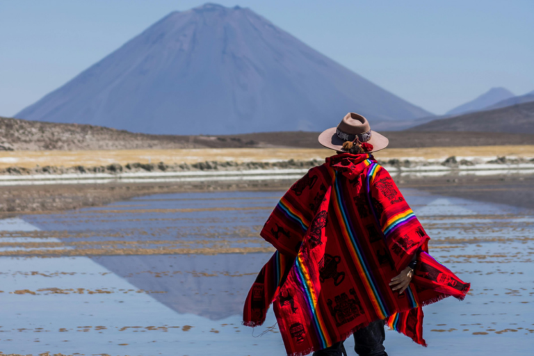 Vista de espaldas de una persona con sombrero marrón y un poncho andino de color rojo vibrante con detalles multicolores, parada frente a la Laguna de Salinas donde se refleja perfectamente la silueta cónica del volcán Ubinas bajo un cielo despejado