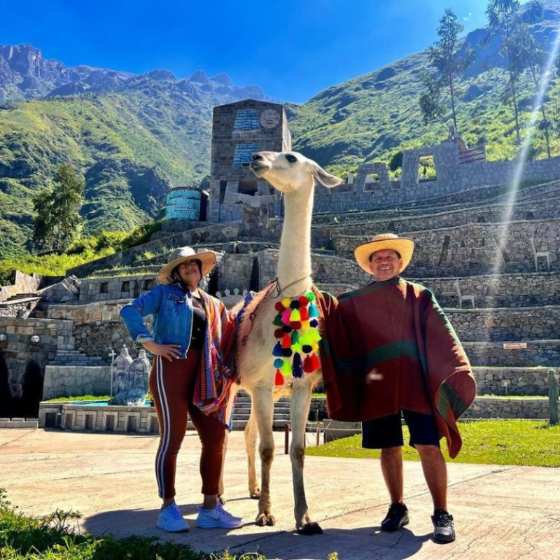 Un hombre y una mujer con sombreros de paja posan sonrientes a ambos lados de una gran llama blanca en un camino de piedra. La llama lleva adornos colgantes de lana con pompones multicolores en el cuello. La mujer a la izquierda viste una chaqueta de jean y leggings marrones, mientras que el hombre a la derecha lleva un poncho tradicional marrón y verde con bermudas negras. Al fondo se aprecian las terrazas y estructuras de piedra del "Machu Picchu Limeño" bajo montañas verdes y un cielo azul brillante.