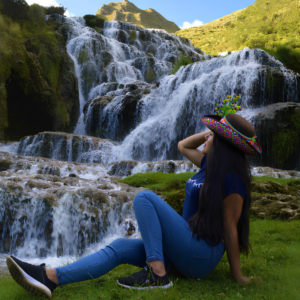 Una joven con jeans y un sombrero tradicional andino decorado con bordados coloridos y flores posa sentada de perfil sobre el pasto, con una hermosa e imponente cascada escalonada de múltiples caídas de agua de fondo bajo un cielo despejado en Ayacucho, Perú.