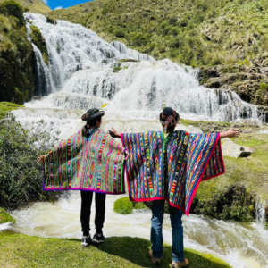 Dos personas de espaldas con los brazos abiertos contemplan una gran cascada escalonada, vistiendo ponchos tradicionales andinos con complejos patrones geométricos multicolores y sombreros típicos de Sarhua en Ayacucho, Perú.
