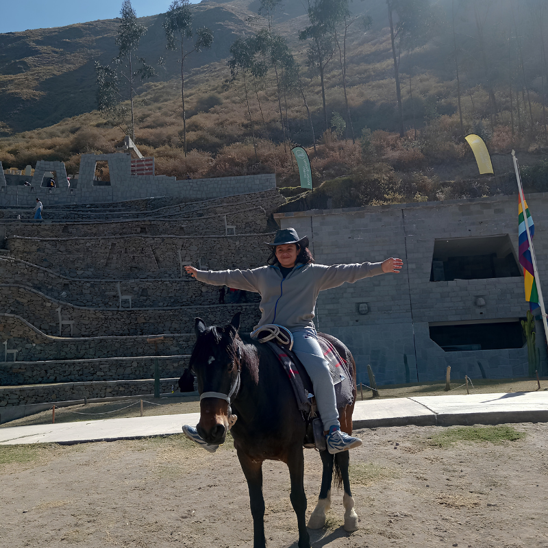 Una persona sonríe montando un caballo marrón oscuro con los brazos abiertos en señal de libertad. Lleva una casaca gris, pantalón deportivo claro y un sombrero de vaquero negro. El caballo se encuentra en una explanada de tierra frente a una imponente construcción de piedra que recrea las terrazas y andenes incas de Machu Picchu, con laderas áridas y árboles de eucalipto al fondo bajo la luz del sol.