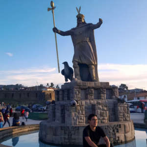 Estatua de bronce del Inca Pachacútec sosteniendo un cetro dorado junto a un halcón de piedra en el centro de una fuente de agua mientras un turista posa sentado en la base en la plaza principal de Vilcashuamán Ayacucho