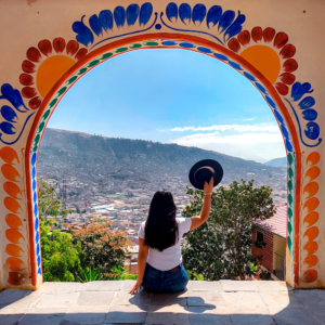 Una mujer sentada de espaldas contempla la ciudad de Ayacucho a través de un arco colonial decorado con flores pintadas a mano, sosteniendo un sombrero negro en el aire.