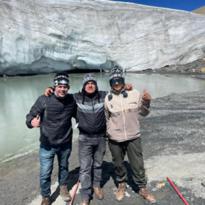 Tres hombres con ropa de abrigo de invierno, botas de montaña y chullos tejidos tradicionales con la palabra "Cusco" posan juntos sonriendo frente a una enorme pared de hielo blanco y gris del Glaciar Quelccaya. Detrás de ellos se observa una laguna de agua de deshielo de color claro y un terreno rocoso y gris de alta montaña.
