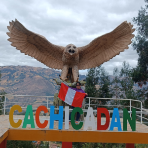Una turista sonriente sostiene la bandera de Perú frente a las letras coloridas de "Cachicadán", debajo de una imponente escultura de un águila con las alas extendidas en un mirador de montaña