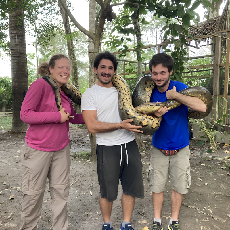 Tres turistas sonríen emocionados mientras cargan juntos una gran serpiente anaconda sobre sus hombros y brazos en un paradero turístico de la selva. Al fondo se observan árboles tropicales y una estructura rústica de madera