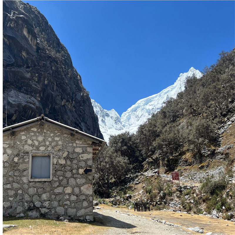 Cabaña rústica con paredes de piedra vista y techo metálico en primer plano, ubicada en un valle altoandino rodeado de árboles nativos de queñua y con un imponente nevado con glaciares bajo un cielo azul despejado al fondo.