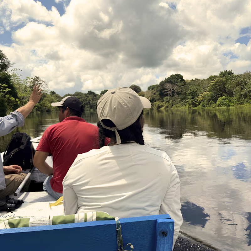 Vista desde la parte trasera de un bote de madera donde varios turistas navegan por un tranquilo río amazónico. Se observa a una mujer de espaldas con gorra beige contemplando el paisaje, rodeada de una densa selva verde bajo un cielo cubierto de nubes que se reflejan en el agua