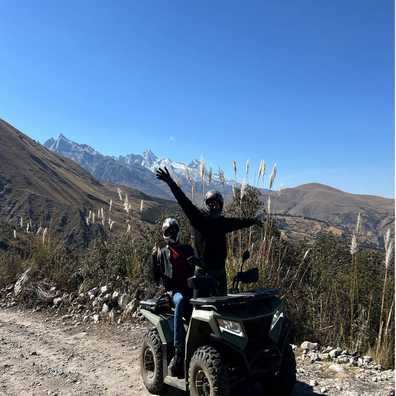 Dos turistas con cascos protectores posan alegres sobre una cuatrimoto verde olivo en un camino de tierra, con una imponente cadena de montañas con picos nevados y cielo azul despejado al fondo