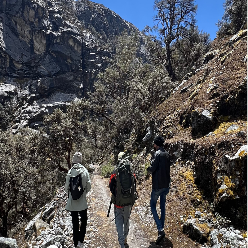 Tres excursionistas con mochilas de montaña y ropa de abrigo caminan de espaldas por un sendero angosto y rocoso, rodeados de laderas empinadas con vegetación nativa y una imponente pared de roca gris al fondo