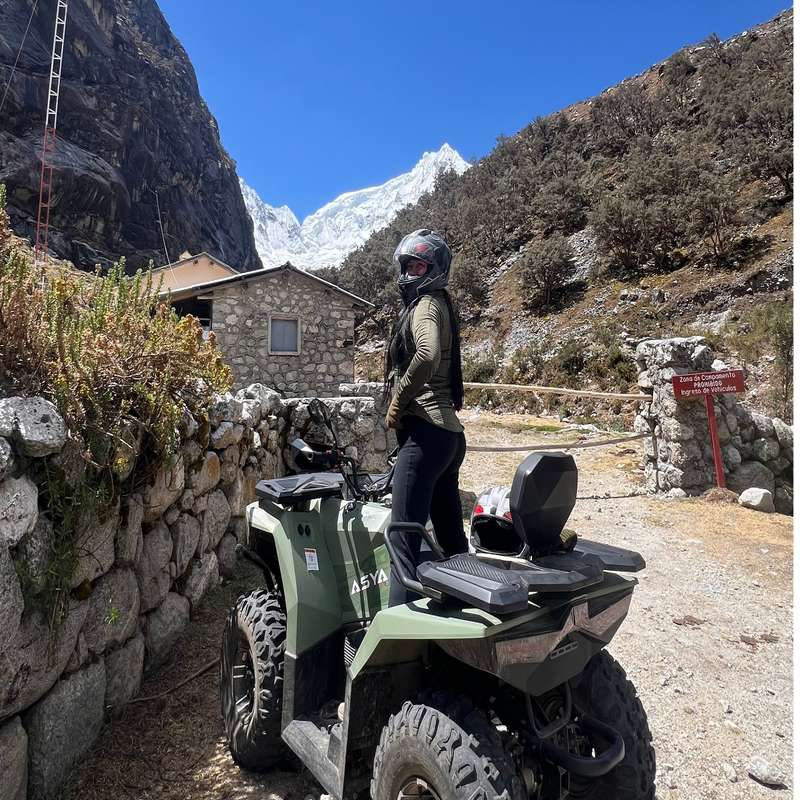Una turista con casco integral posa de pie sobre una cuatrimoto verde oliva junto a un muro de piedra artesanal, mostrando al fondo una cabaña rústica y un imponente nevado con glaciares bajo el cielo azul