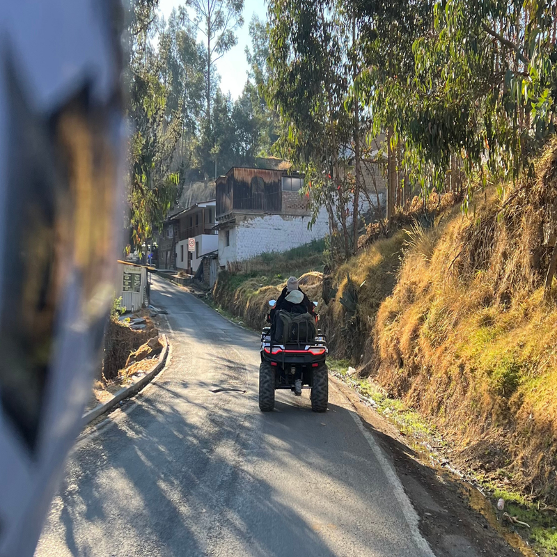 Vista desde atrás de una cuatrimoto todoterreno con dos pasajeros avanzando por una carretera asfaltada cuesta arriba, flanqueada por laderas con vegetación, árboles de eucalipto y casas rústicas al fondo