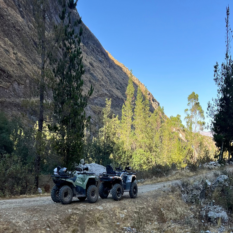 Dos cuatrimotos todoterreno estacionadas en un camino de tierra rústico, rodeadas de pinos altos y laderas empinadas bajo un cielo azul despejado durante una excursión de aventura
