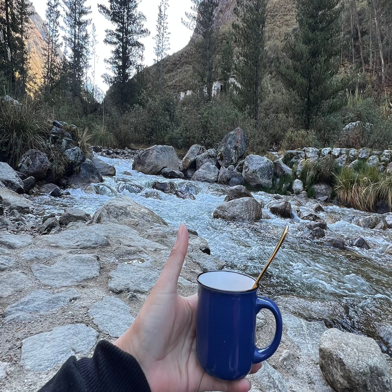 Una mano sostiene una taza azul con una cuchara de metal dorada en primer plano, con un río de agua cristalina y rápida corriendo entre grandes rocas y árboles altos de fondo en un paisaje de montaña