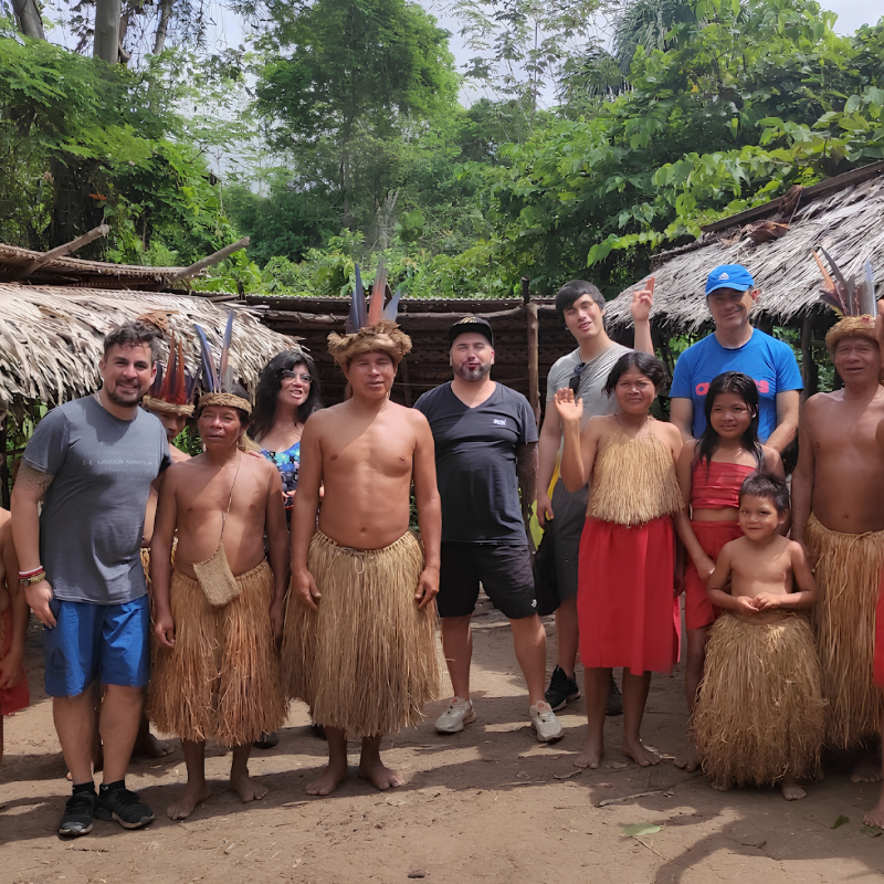 Un grupo de turistas posa sonriente junto a hombres, mujeres y niños de una comunidad nativa amazónica en su aldea. Los miembros locales visten trajes tradicionales con faldas de fibra de paja, collares y coloridos tocados de plumas en la cabeza, rodeados de malocas rústicas y la vegetación de la selva.