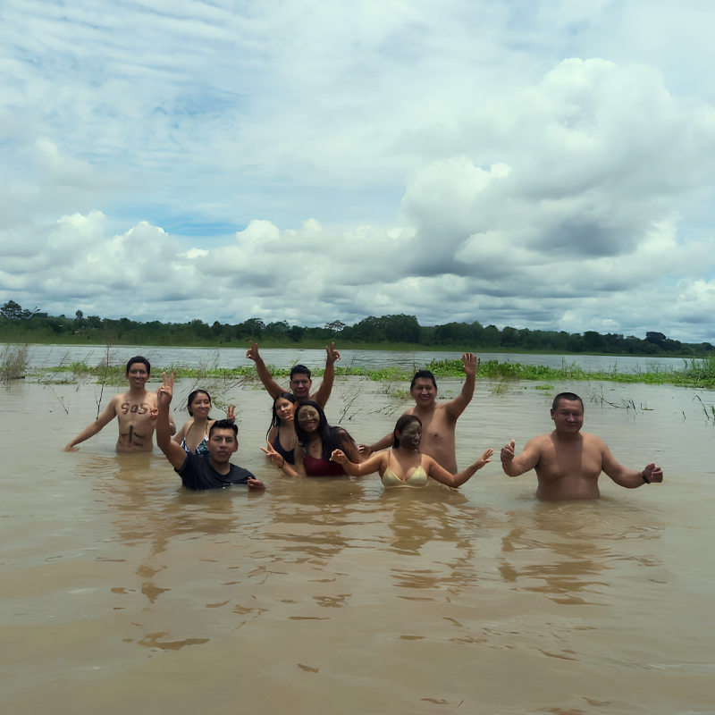 Un grupo de turistas sonríe y saluda con los brazos en alto mientras se sumerge en las aguas de un ancho río amazónico. Al fondo se divisa la línea de vegetación de la selva bajo un cielo nublado, destacando la experiencia de aventura en un tour de varios días