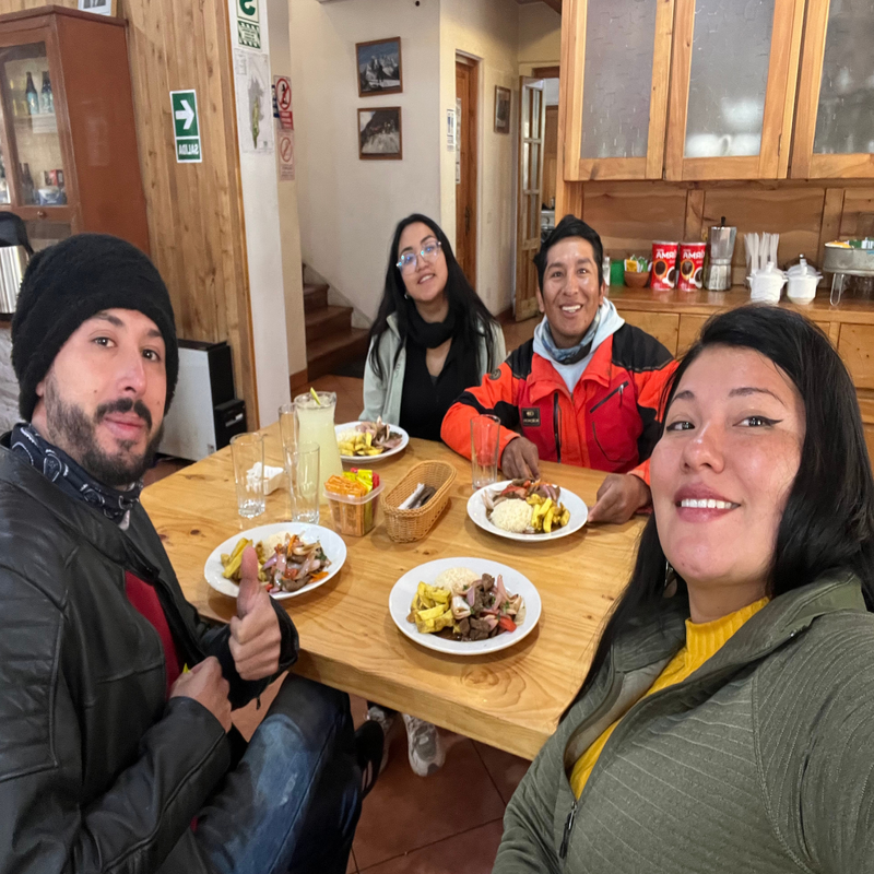 Cuatro turistas sonríen sentados alrededor de una mesa de madera rústica disfrutando de platos individuales de lomo saltado peruano con arroz y papas fritas, acompañados de una jarra de limonada en un comedor cálido