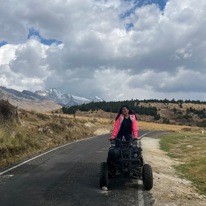Mujer con casaca rosada sonríe manejando una cuatrimoto en una carretera asfaltada con montañas y nevados de fondo en Huaraz